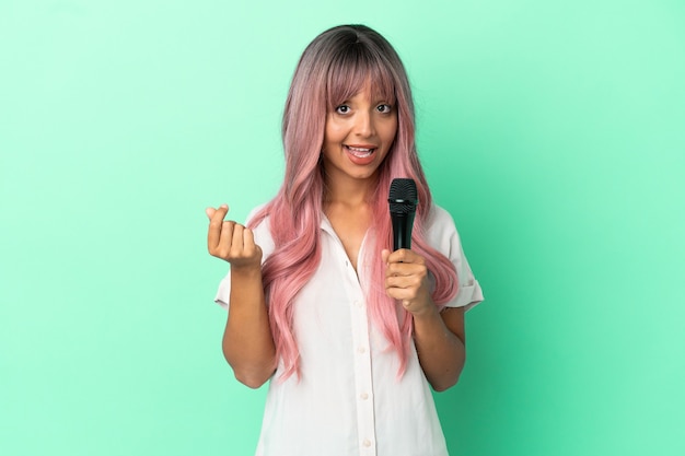 Joven mujer cantante de raza mixta con cabello rosado aislado sobre fondo verde haciendo gesto de dinero