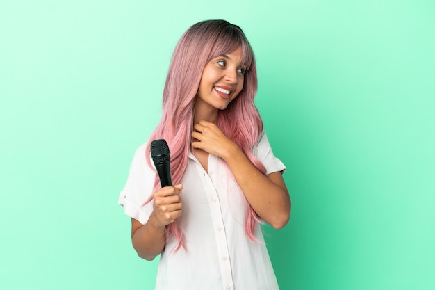 Joven mujer cantante de raza mixta con cabello rosado aislado sobre fondo verde mirando hacia arriba mientras sonríe