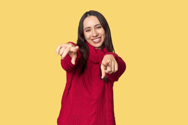 Foto joven mujer caucásica con un acogedor suéter rojo en el fondo del estudio sonríe alegremente señalando hacia el frente