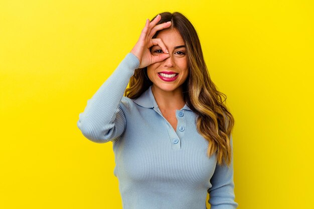 Joven mujer caucásica aislada en amarillo emocionado manteniendo el gesto ok en el ojo.