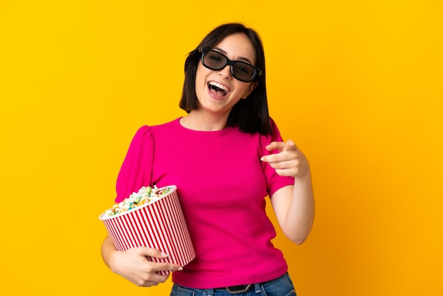 Joven mujer caucásica aislada en amarillo con gafas 3d y sosteniendo un gran cubo de palomitas de maíz mientras apunta al frente