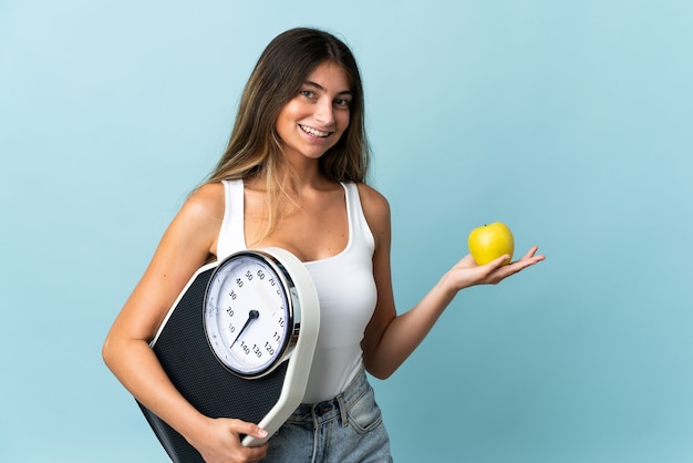 Joven mujer caucásica aislada en azul con báscula y con una manzana