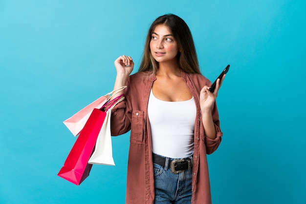 Joven mujer caucásica aislada en azul sosteniendo bolsas de la compra y un teléfono móvil