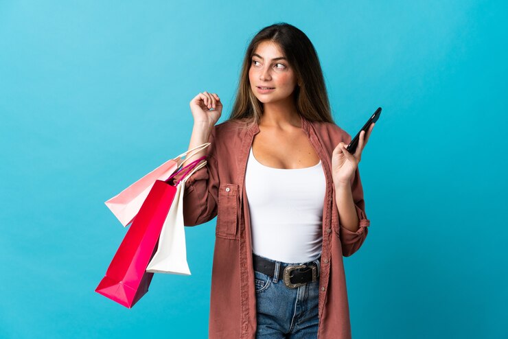 Foto joven mujer caucásica aislada en azul sosteniendo bolsas de la compra y un teléfono móvil