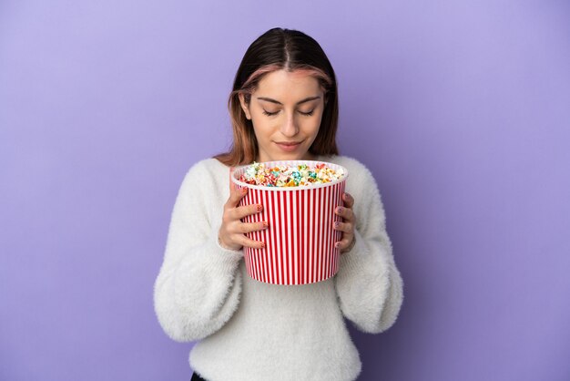 Joven mujer caucásica aislada en azul sosteniendo un gran balde de palomitas de maíz