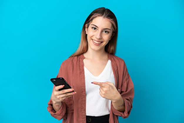 Joven mujer caucásica aislada en azul mediante teléfono móvil y apuntando