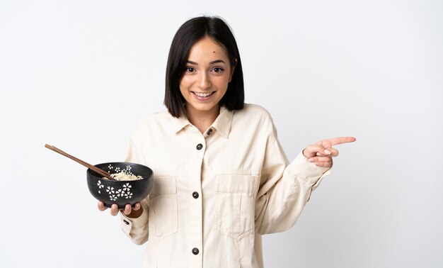 Joven mujer caucásica aislada en blanco sorprendida y apuntando con el dedo hacia un lado mientras sostiene un plato de fideos con palillos