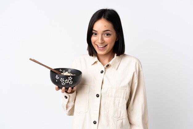 Joven mujer caucásica aislada en blanco con sorpresa y expresión facial conmocionada mientras sostiene un plato de fideos con palillos