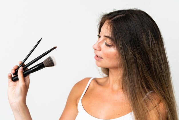 Joven mujer caucásica aislada en blanco sosteniendo pincel de maquillaje y mirando