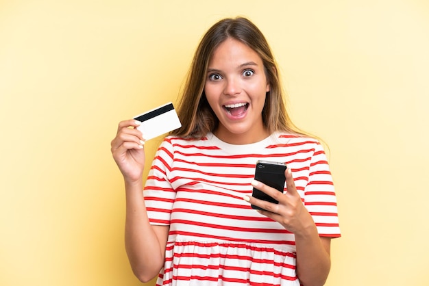 Joven mujer caucásica aislada de fondo amarillo comprando con el móvil y sosteniendo una tarjeta de crédito con expresión sorprendida