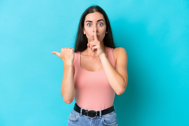 Joven mujer caucásica aislada de fondo azul apuntando hacia un lado y haciendo gesto de silencio