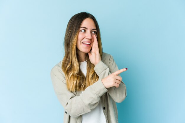 Joven mujer caucásica aislada de fondo azul diciendo un chisme, señalando a un lado informando algo.