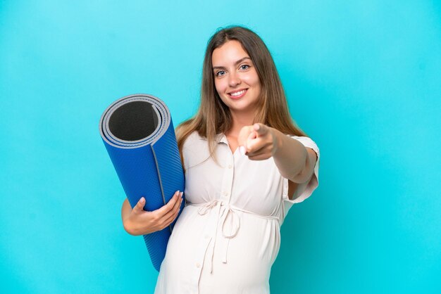 Joven mujer caucásica aislada de fondo azul embarazada y yendo a clases de yoga mientras apunta al frente