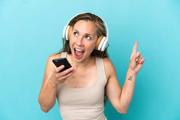 Joven mujer caucásica aislada de fondo azul escuchando música con un móvil y cantando