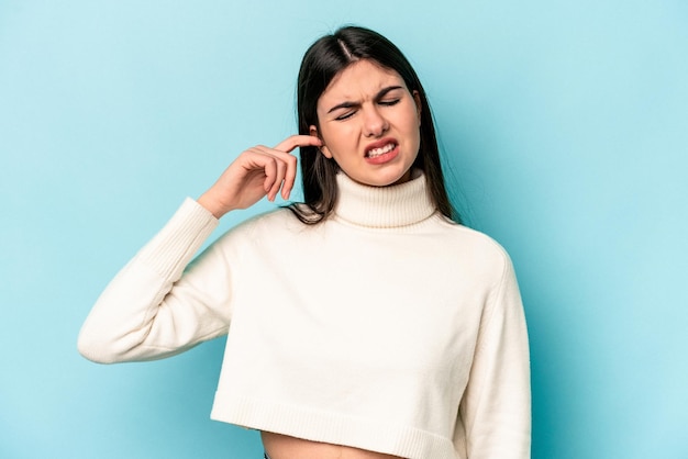 Joven mujer caucásica aislada de fondo azul que cubre las orejas con los dedos estresada y desesperada por un ambiente ruidoso