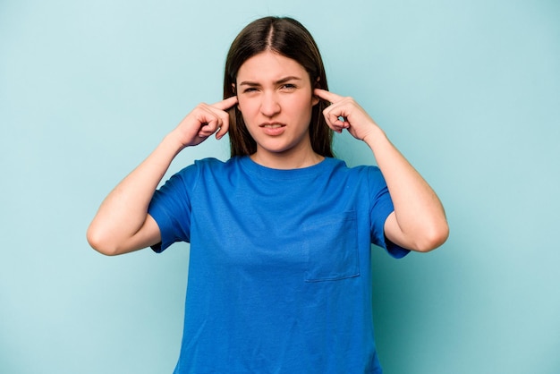 Joven mujer caucásica aislada de fondo azul que cubre las orejas con los dedos estresada y desesperada por un ambiente ruidoso