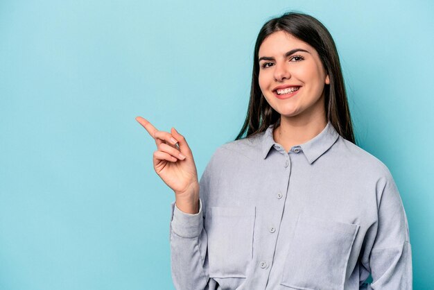 Joven mujer caucásica aislada de fondo azul sonriendo alegremente señalando con el dedo índice lejos