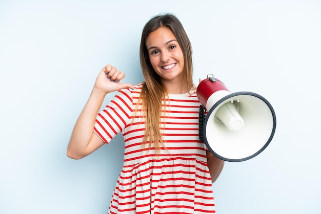 Joven mujer caucásica aislada de fondo azul sosteniendo un megáfono y orgullosa y satisfecha