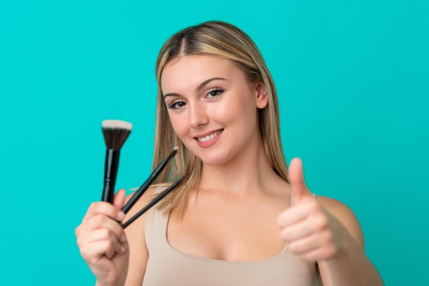 Joven mujer caucásica aislada de fondo azul sosteniendo un pincel de maquillaje y con el pulgar hacia arriba