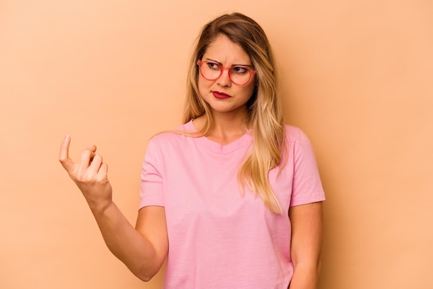 Joven mujer caucásica aislada de fondo beige apuntándote con el dedo como si invitara a acercarse