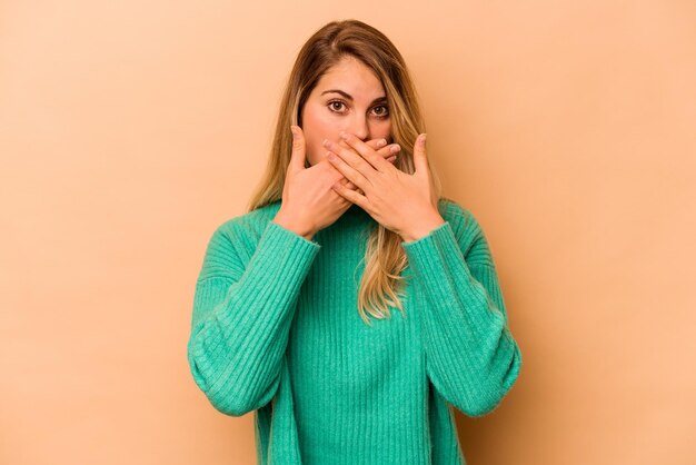 Joven mujer caucásica aislada de fondo beige cubriendo la boca con las manos preocupadas