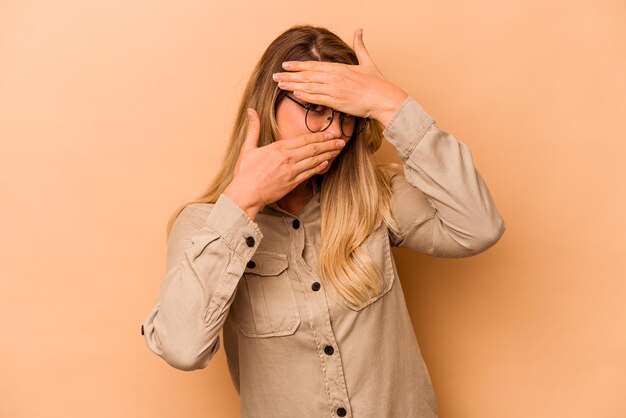 Joven mujer caucásica aislada de fondo beige parpadea a la cámara a través de los dedos avergonzada cubriendo la cara