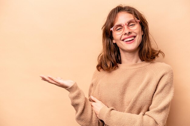 Joven mujer caucásica aislada de fondo beige que muestra un espacio de copia en una palma y sostiene otra mano en la cintura