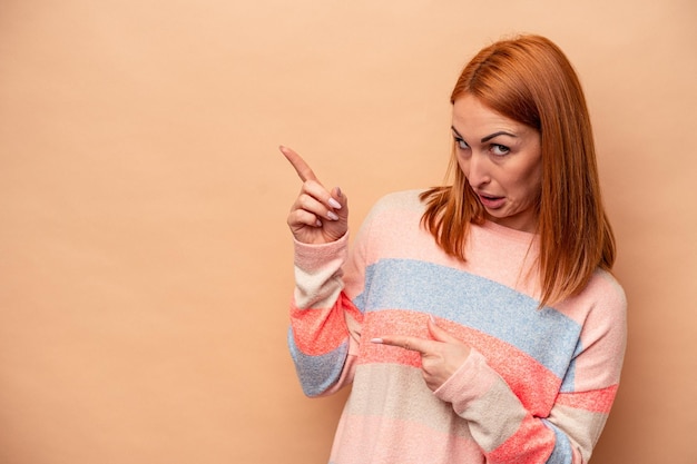 Joven mujer caucásica aislada de fondo beige señalando con los dedos índices a un espacio de copia que expresa entusiasmo y deseo