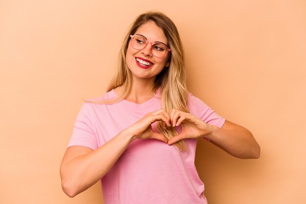 Joven mujer caucásica aislada de fondo beige sonriendo y mostrando una forma de corazón con las manos