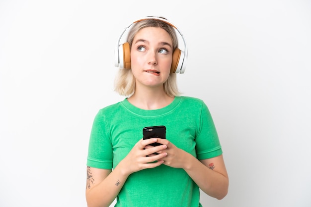 Joven mujer caucásica aislada de fondo blanco escuchando música con un móvil y pensando