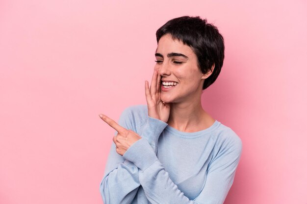 Joven mujer caucásica aislada de fondo rosa diciendo un chisme apuntando a un lado informando algo