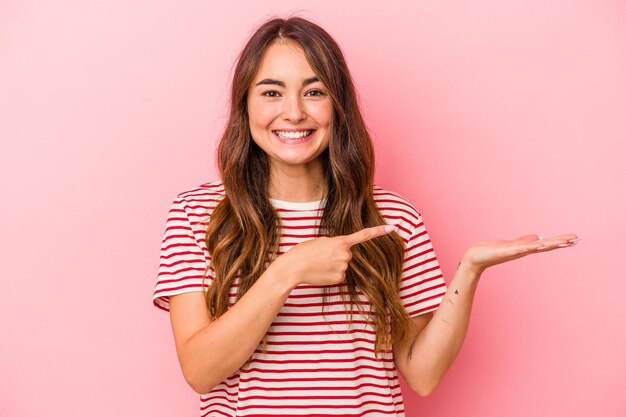 Joven mujer caucásica aislada de fondo rosa emocionada sosteniendo un espacio de copia en la palma.