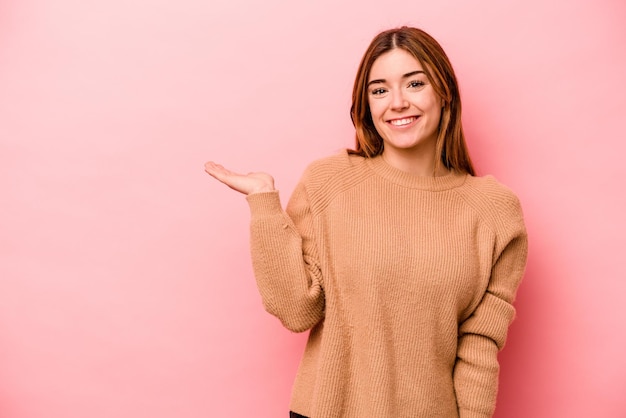 Joven mujer caucásica aislada de fondo rosa mostrando un espacio de copia en una palma y sosteniendo otra mano en la cintura