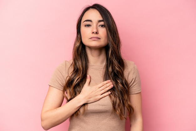 Joven mujer caucásica aislada de fondo rosa prestando juramento poniendo la mano en el pecho