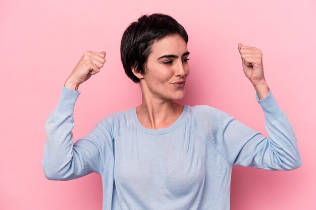 Joven mujer caucásica aislada de fondo rosa que muestra un gesto de fuerza con los brazos, símbolo del poder femenino