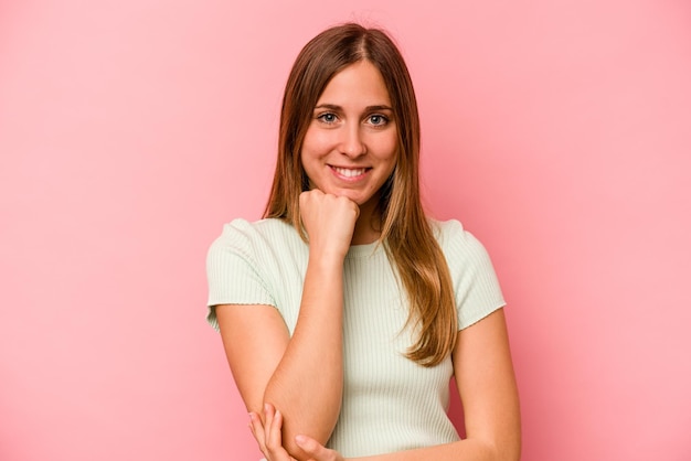 Joven mujer caucásica aislada de fondo rosa sonriendo feliz y confiada tocando la barbilla con la mano