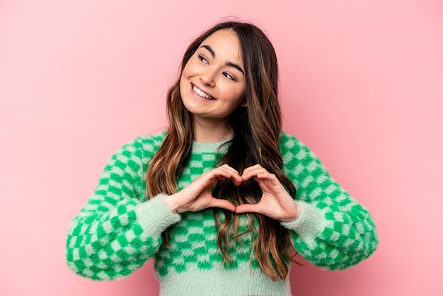 Joven mujer caucásica aislada de fondo rosa sonriendo y mostrando una forma de corazón con las manos