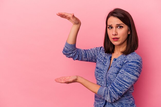 Joven mujer caucásica aislada de fondo rosa sorprendida y asombrada sosteniendo un espacio de copia entre las manos