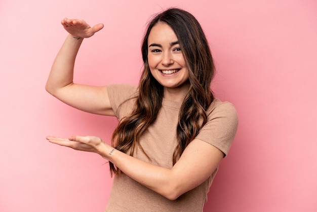 Joven mujer caucásica aislada de fondo rosa sosteniendo algo pequeño con los dedos índices sonriendo y confiada