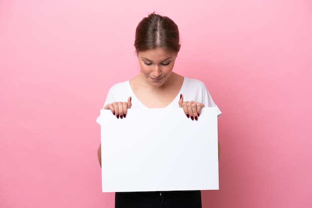 Joven mujer caucásica aislada de fondo rosa sosteniendo un cartel vacío y mirándolo