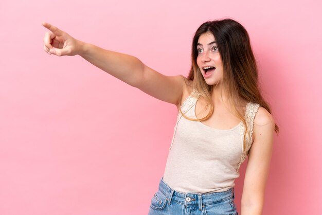 Joven mujer caucásica aislada en un fondo rosado señalando hacia otro lado