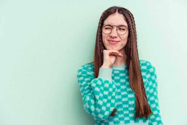 Joven mujer caucásica aislada de fondo verde tocando la parte posterior de la cabeza pensando y tomando una decisión