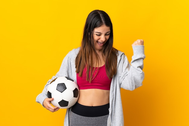 Joven mujer caucásica aislada en la pared amarilla con balón de fútbol celebrando una victoria