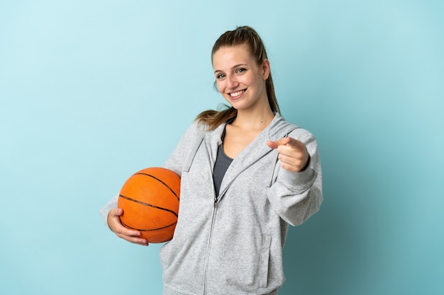 Joven mujer caucásica aislada en la pared azul jugando baloncesto y apuntando hacia el frente