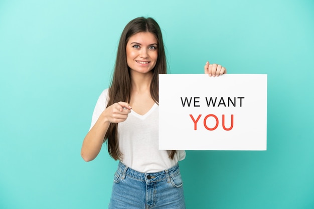 Joven mujer caucásica aislada en la pared azul sosteniendo We Want You board y apuntando hacia el frente