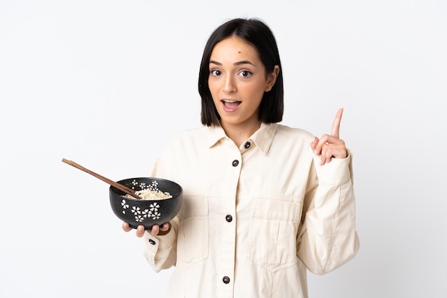 Joven mujer caucásica aislada en la pared blanca apuntando hacia una gran idea mientras sostiene un plato de fideos con palillos