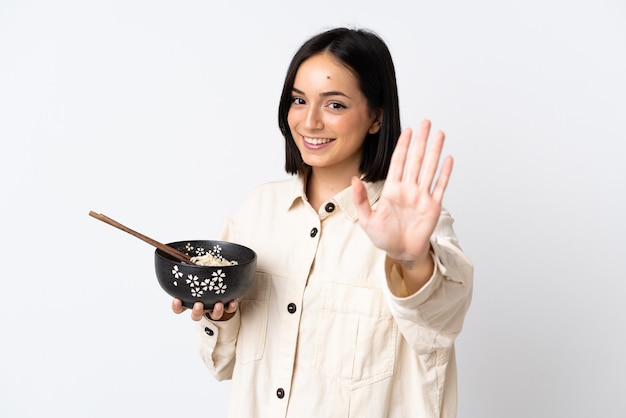 Joven mujer caucásica aislada en la pared blanca saludando con la mano con expresión feliz mientras sostiene un plato de fideos con palillos