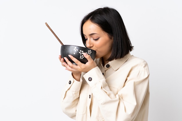 Joven mujer caucásica aislada en la pared blanca sosteniendo un plato de fideos con palillos y comiéndolo