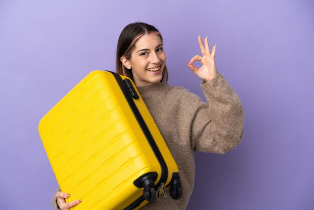 Joven mujer caucásica aislada en la pared púrpura en vacaciones con maleta de viaje y haciendo el signo de OK