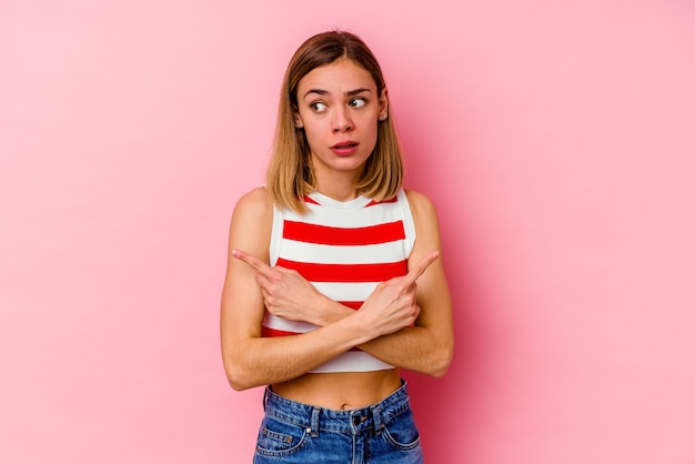 Joven mujer caucásica aislada en la pared rosa apunta hacia los lados, está tratando de elegir entre dos opciones.